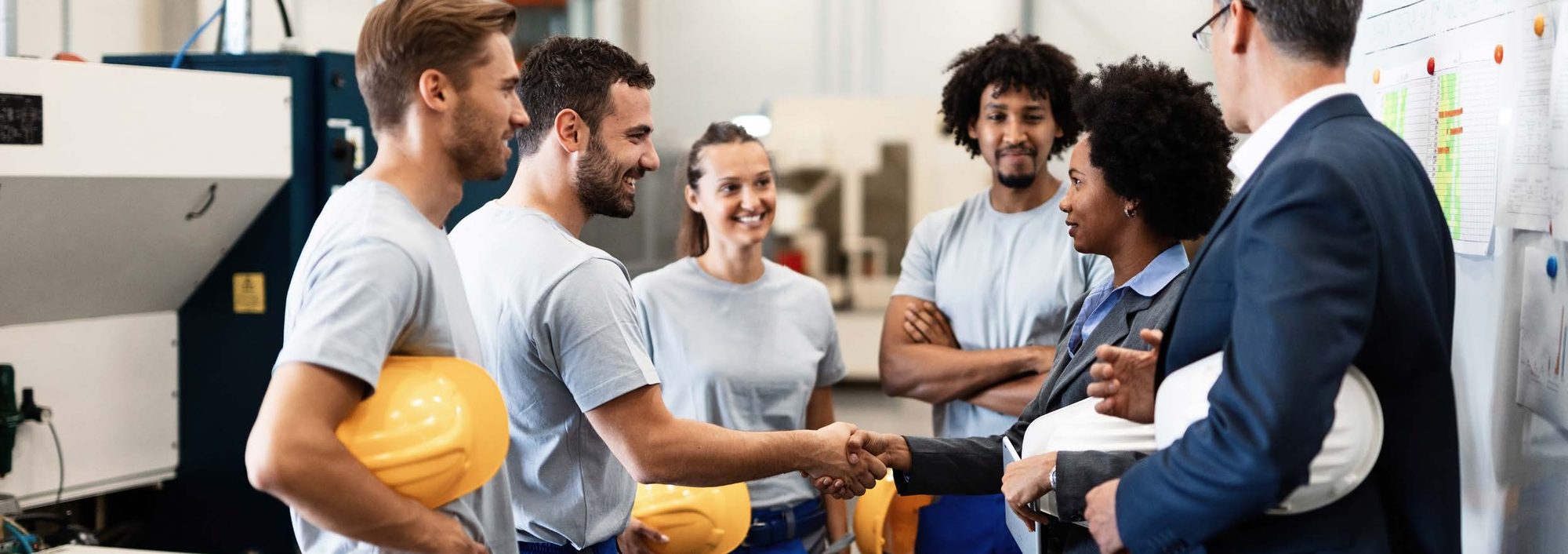 A team of factory workers shake hands with colleagues in suits, gathered in a workshop